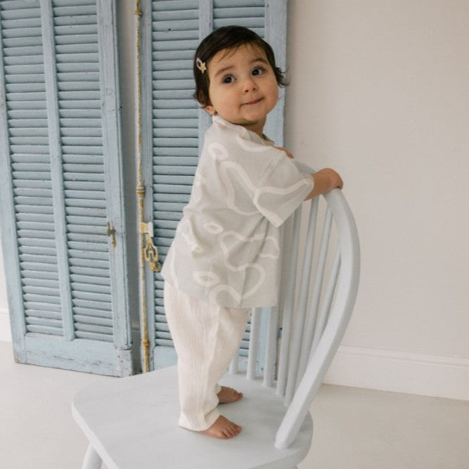 Child in a white pear t-shirt standing on a white chair with light blue shutters in the background